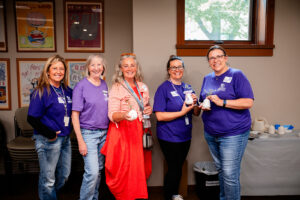Five women stand near one another, three holding small vases of flowers, while all smiling