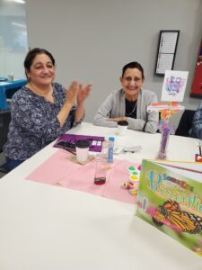 Two people sitting at a table with craft items and a book, smiling and interacting.