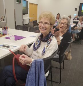 The image depicts a group of people seated at a long table in a classroom or meeting room setting. In the foreground, a person is smiling and sitting at the edge of the table, wearing a white sweater and a colorful scarf.