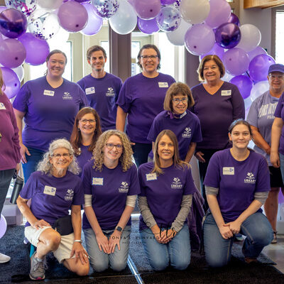 A group of LMP members posing together all wearing purple in front of a balloon arch