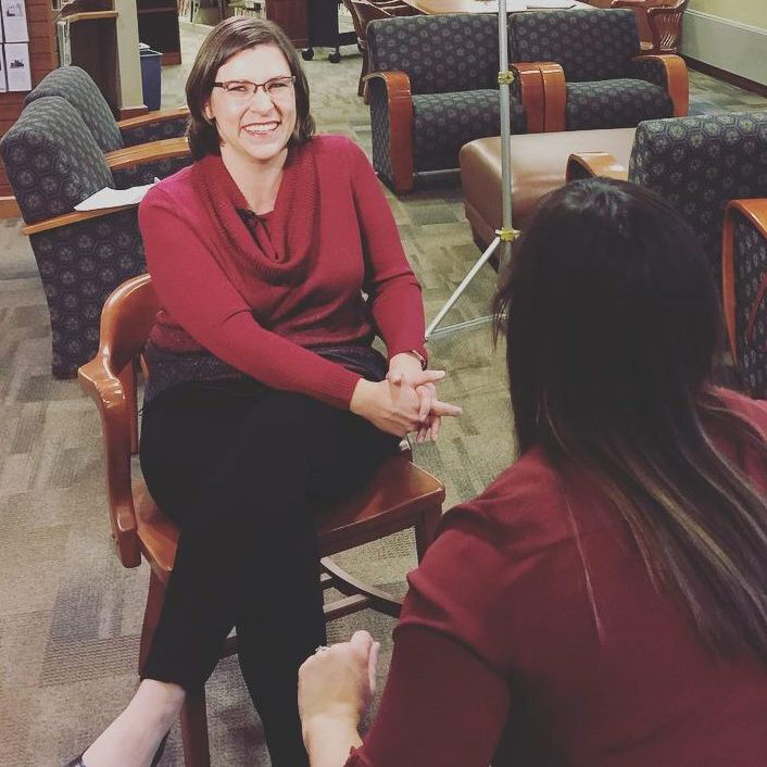 A woman sitting in a chair in a lobby being interviewed by another woman